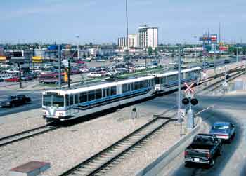 Twin-car Calgary LRV trainset using dual carriageway median crosses level crossing protected by flashing lights and half barriers.